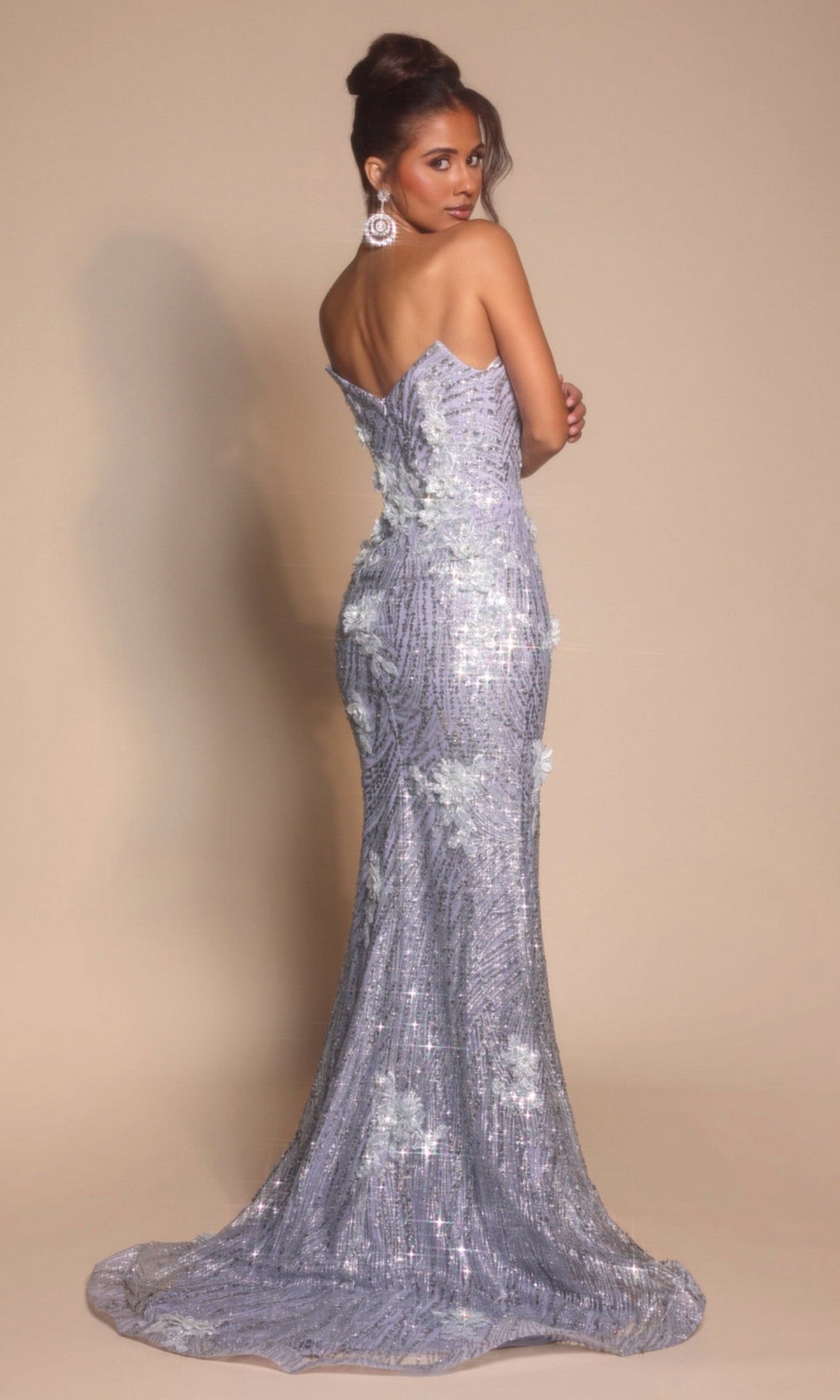 A woman faces backward in the Portia and Scarlett PS26221, a floor-length lavender prom dress with floral details and sparkles, standing against a plain beige background.